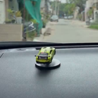 A small lime green toy sports car mounted on a black circular suction cup base, placed on a dark car dashboard with a blurred street scene visible through the windshield.