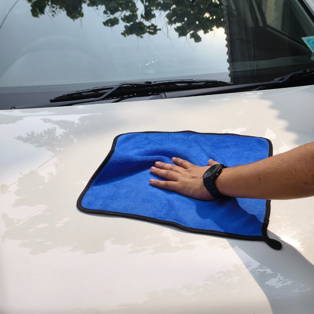 Person cleaning a car with a blue microfiber towel on a sunny day.